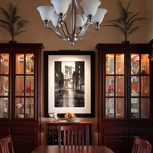 Black-and-white framed artwork illuminated by a Plug-in Vision Series art light in an elegant dining room with dark wood cabinets and decor.