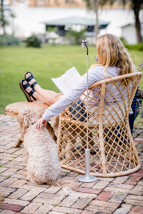 Woman reading outdoors comfortably seated in a wicker chair, using the portable LightBob™: a cordless, go-anywhere LED task floor light, providing focused illumination on her book.